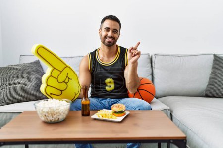 Photo for Young hispanic man with beard holding basketball ball cheering tv game smiling happy pointing with hand and finger to the side - Royalty Free Image