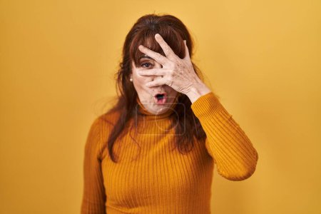 Photo for Middle age hispanic woman standing over yellow background peeking in shock covering face and eyes with hand, looking through fingers with embarrassed expression. - Royalty Free Image