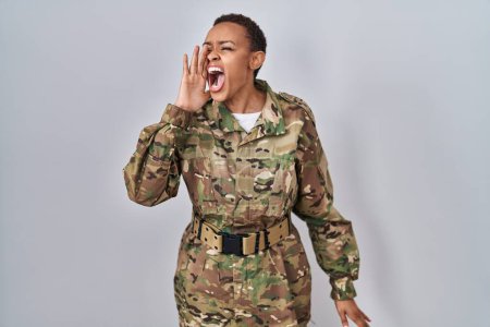 Foto de Hermosa mujer afroamericana vistiendo camuflaje uniforme del ejército gritando y gritando fuerte a un lado con la mano en la boca. concepto de comunicación. - Imagen libre de derechos