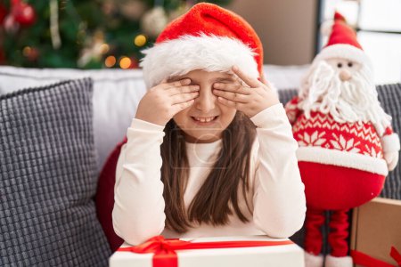 Foto de Adorable chica hispana sentada en el sofá por la decoración de Navidad con las manos en los ojos en casa - Imagen libre de derechos