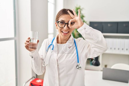 Foto de Joven morena médico mujer sosteniendo vaso de agua sonriendo feliz haciendo ok signo con la mano en el ojo mirando a través de los dedos - Imagen libre de derechos