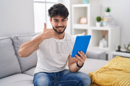 Photo for Hispanic man with beard using touchpad sitting on the sofa smiling doing phone gesture with hand and fingers like talking on the telephone. communicating concepts. - Royalty Free Image
