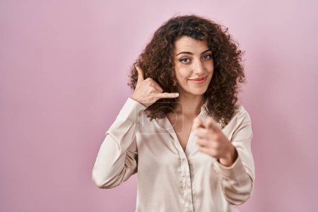 Téléchargez les photos : Femme hispanique avec les cheveux bouclés debout sur fond rose souriant faire parler au téléphone geste et vous pointant du doigt. Appelle-moi. - en image libre de droit