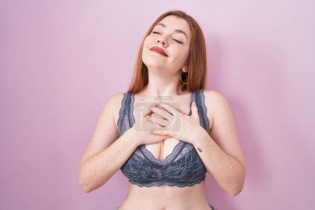 Foto de Redhead woman wearing lingerie over pink background smiling with hands on chest with closed eyes and grateful gesture on face. health concept. - Imagen libre de derechos