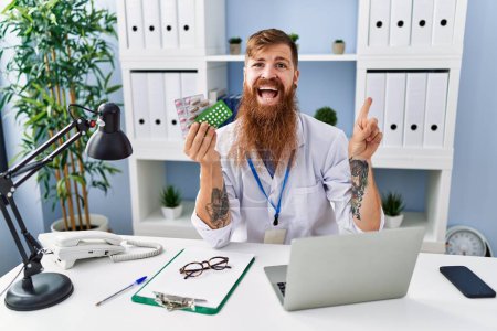 Téléchargez les photos : Homme rousse à longue barbe portant l'uniforme de médecin tenant des pilules sur ordonnance souriant heureux pointant avec la main et le doigt sur le côté - en image libre de droit
