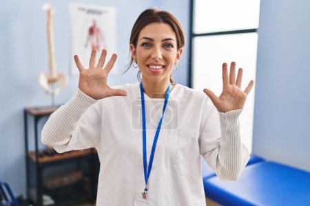 Foto de Joven morena que trabaja en la clínica de recuperación del dolor mostrando y señalando con los dedos número diez mientras sonríe confiado y feliz. - Imagen libre de derechos