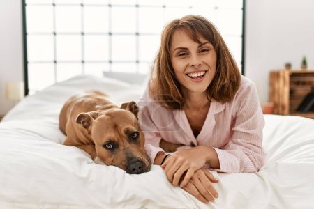 Téléchargez les photos : Jeune femme caucasienne souriante confiante couchée sur le lit avec chien dans la chambre - en image libre de droit