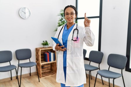 Téléchargez les photos : Jeune femme médecin afro-américaine dans la salle d'attente surpris par une idée ou une question pointant du doigt avec un visage heureux, numéro un - en image libre de droit