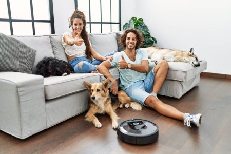 Téléchargez les photos : Jeune couple hispanique relaxant à la maison par aspirateur robot souriant heureux et positif, pouce levé faire excellent et signe d'approbation - en image libre de droit