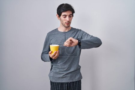 Téléchargez les photos : Jeune homme hispanique portant un pyjama boire une tasse de café en regardant l'heure de la montre inquiet, peur de se retrouver en retard - en image libre de droit