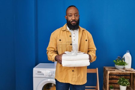 Téléchargez les photos : African american man holding clean towels at laundry room relaxed with serious expression on face. simple and natural looking at the camera. - en image libre de droit