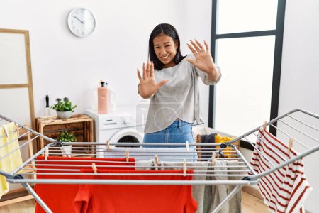 Foto de Young hispanic woman putting fresh laundry on clothesline afraid and terrified with fear expression stop gesture with hands, shouting in shock. panic concept. - Imagen libre de derechos