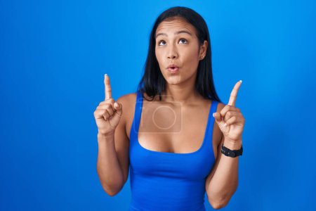 Photo for Hispanic woman standing over blue background amazed and surprised looking up and pointing with fingers and raised arms. - Royalty Free Image