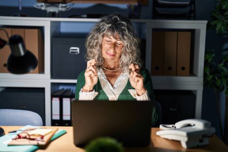 Foto de Middle age woman working at night using computer laptop gesturing finger crossed smiling with hope and eyes closed. luck and superstitious concept. - Imagen libre de derechos