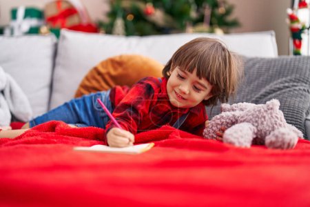 Téléchargez les photos : Adorable tout-petit écrivant sur un cahier couché sur un canapé près d'un arbre de Noël à la maison - en image libre de droit