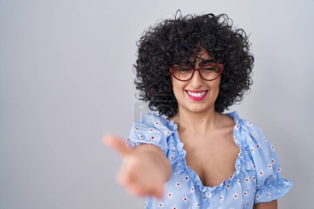 Photo for Young brunette woman with curly hair wearing glasses over isolated background smiling friendly offering handshake as greeting and welcoming. successful business. - Royalty Free Image