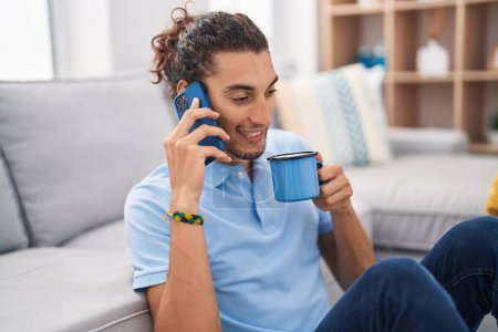 Photo for Young hispanic man talking on smartphone drinking coffee at home - Royalty Free Image