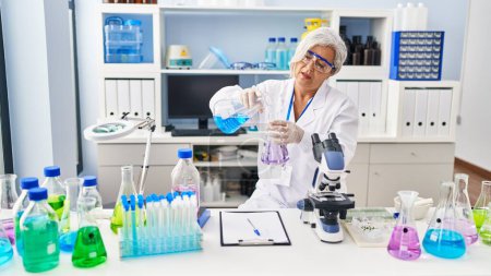 Foto de Middle age woman wearing scientist uniform pouring liquid on test tube at laboratory - Imagen libre de derechos