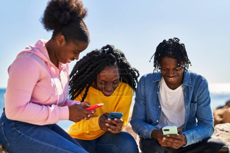 Foto de Amigos afroamericanos usando teléfono inteligente sentado en la roca en la playa - Imagen libre de derechos