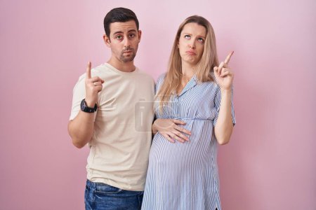 Photo for Young couple expecting a baby standing over pink background pointing up looking sad and upset, indicating direction with fingers, unhappy and depressed. - Royalty Free Image