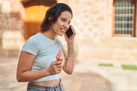 Foto de Young hispanic woman talking on the smartphone eating ice cream at street - Imagen libre de derechos