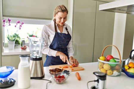 Foto de Joven rubia sonriendo confiada cortando zanahoria en la cocina - Imagen libre de derechos