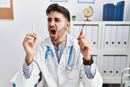 Photo for Young doctor man holding electronic cigarette at medical clinic angry and mad screaming frustrated and furious, shouting with anger. rage and aggressive concept. - Royalty Free Image