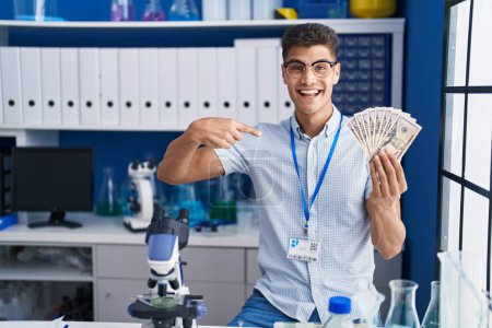 Foto de Joven hispano trabajando en laboratorio científico sosteniendo dólares apuntando con el dedo a uno mismo sonriendo feliz y orgulloso - Imagen libre de derechos