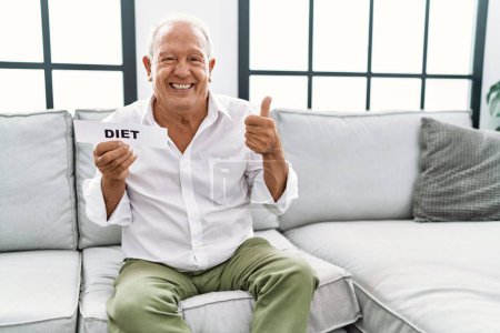 Foto de Hombre mayor sosteniendo banner de dieta sonriendo feliz y positivo, pulgar hacia arriba haciendo excelente y signo de aprobación - Imagen libre de derechos