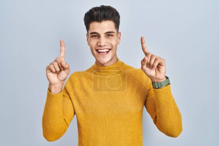 Photo for Young hispanic man standing over blue background smiling amazed and surprised and pointing up with fingers and raised arms. - Royalty Free Image
