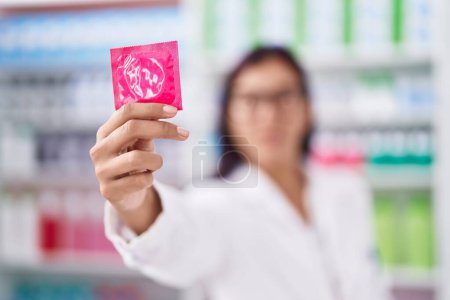 Téléchargez les photos : Jeune belle hispanique femme pharmacien souriant confiant tenant préservatif à la pharmacie - en image libre de droit