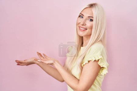 Foto de Caucasian woman standing over pink background inviting to enter smiling natural with open hand - Imagen libre de derechos