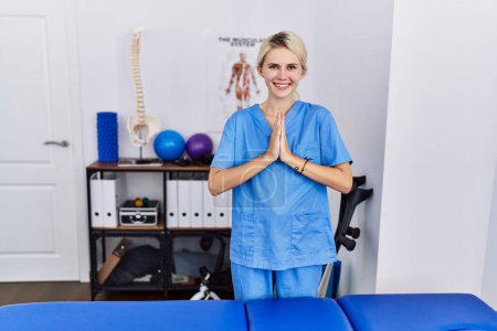 Foto de Joven fisioterapeuta mujer que trabaja en la clínica de recuperación del dolor rezando con las manos juntas pidiendo perdón sonriendo confiado. - Imagen libre de derechos