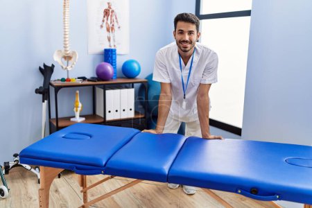 Téléchargez les photos : Young hispanic man wearing physiotherapist uniform leaning on massage table at rehab clinic - en image libre de droit