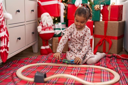 Photo for Adorable hispanic girl playing with train toy sitting on floor by christmas tree at home - Royalty Free Image