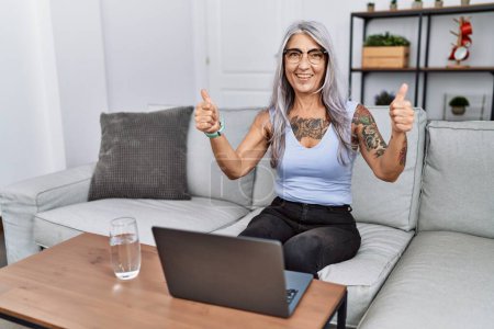 Téléchargez les photos : Femme aux cheveux gris d'âge moyen utilisant un ordinateur portable à la maison signe de succès faire un geste positif avec la main, pouces levés souriant et heureux. expression joyeuse et geste gagnant. - en image libre de droit