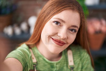 Téléchargez les photos : Jeune rousse femme fleuriste souriant confiant faire selfie par caméra à la boutique de fleurs - en image libre de droit