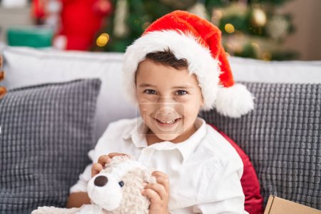 Photo for Adorable hispanic toddler hugging teddy bear sitting on sofa by christmas tree at home - Royalty Free Image