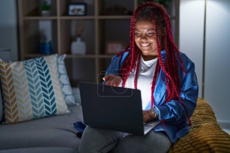 Foto de African american woman with braided hair using computer laptop at night smiling cheerful offering palm hand giving assistance and acceptance. - Imagen libre de derechos