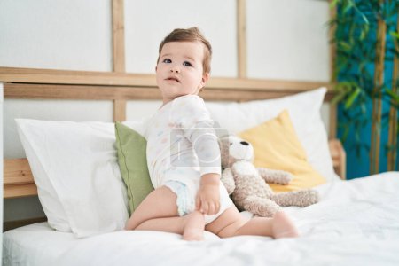 Foto de Adorable bebé caucásico sentado en la cama con expresión relajada en el dormitorio - Imagen libre de derechos
