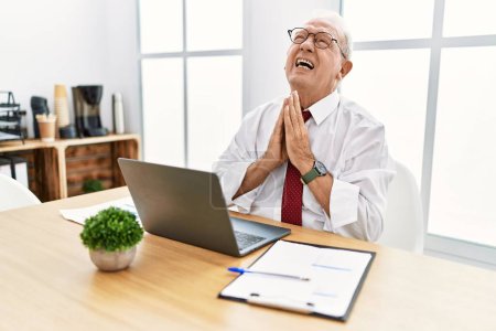 Photo for Senior man working at the office using computer laptop begging and praying with hands together with hope expression on face very emotional and worried. begging. - Royalty Free Image