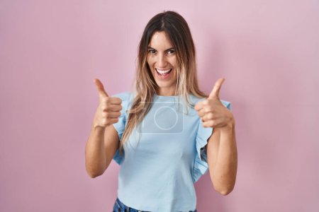 Foto de Mujer hispana joven de pie sobre el signo de éxito de fondo rosa haciendo gesto positivo con la mano, pulgares hacia arriba sonriendo y feliz. expresión alegre y gesto ganador. - Imagen libre de derechos