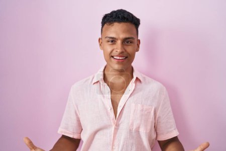 Foto de Joven hombre hispano de pie sobre fondo rosa sonriente alegre con los brazos abiertos como bienvenida amistosa, saludos positivos y confiados - Imagen libre de derechos