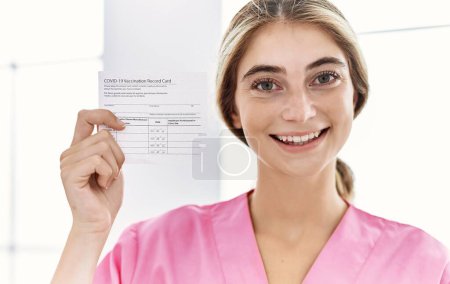 Photo for Young blonde woman holding covid record card looking positive and happy standing and smiling with a confident smile showing teeth - Royalty Free Image