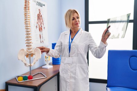 Téléchargez les photos : Femme blonde d'âge moyen portant un uniforme de physiothérapeute tenant des rayons X à la clinique de physiothérapie - en image libre de droit