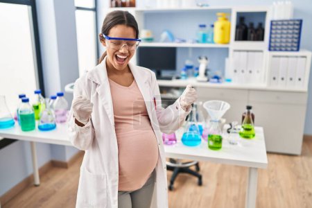 Téléchargez les photos : Jeune femme enceinte travaillant au laboratoire scientifique très heureuse et excitée de faire geste gagnant avec les bras levés, souriant et criant pour le succès. concept de célébration. - en image libre de droit