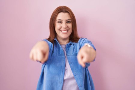 Foto de Mujer hispana joven con el pelo rojo de pie sobre el fondo rosa que apunta a usted y la cámara con los dedos, sonriendo positiva y alegre - Imagen libre de derechos