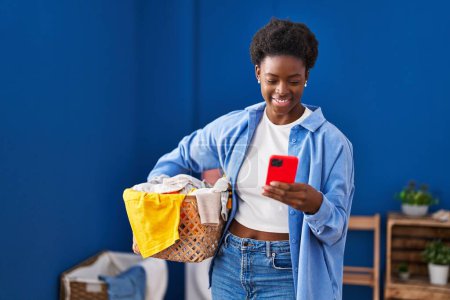 Téléchargez les photos : African american woman holding basket with clothes using smartphone at laundry room - en image libre de droit