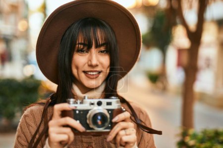 Téléchargez les photos : Brunette femme portant chapeau d'hiver souriant à l'aide d'une caméra vintage en plein air à la ville - en image libre de droit