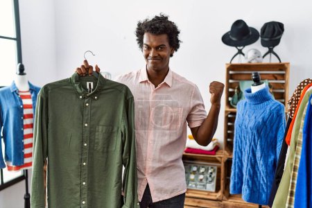 Photo for African man with curly hair holding shirt from clothing rack at retail shop screaming proud, celebrating victory and success very excited with raised arms - Royalty Free Image